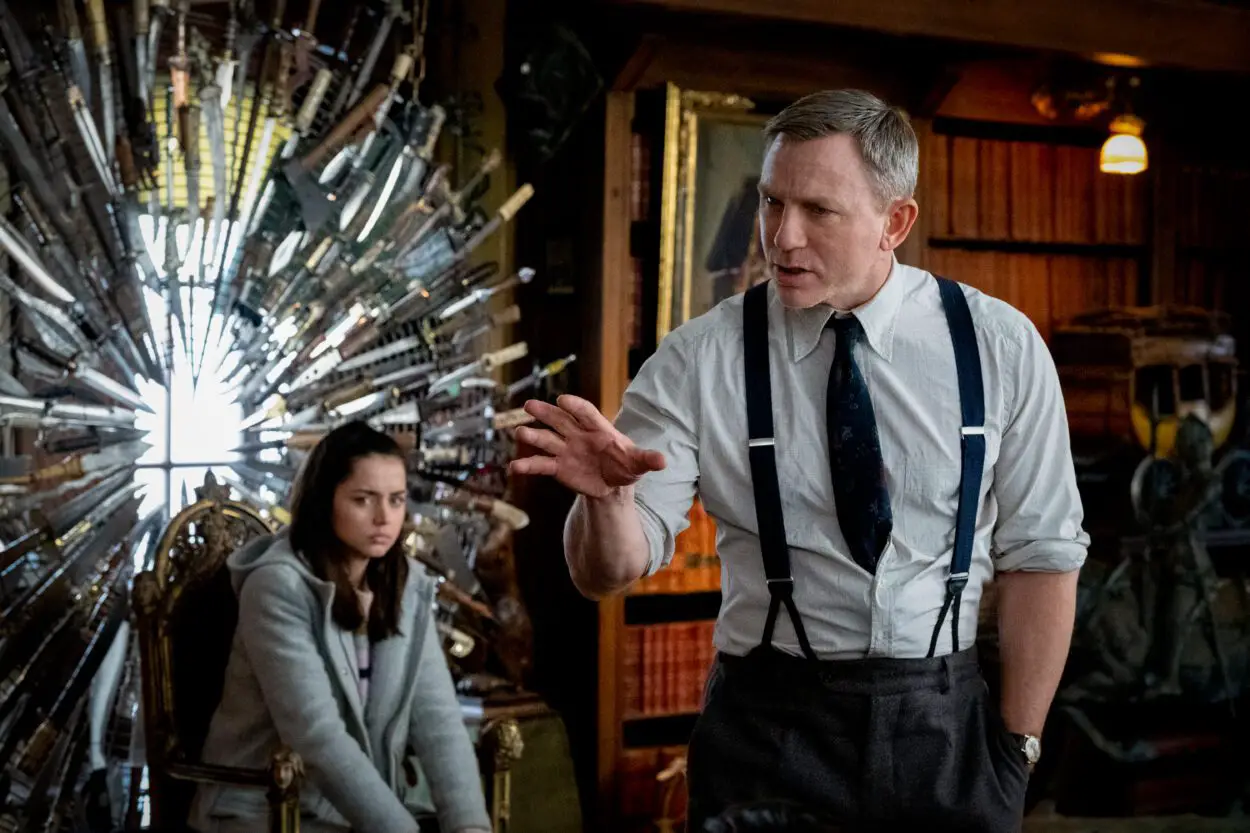 Detective Benoit Blanc (Daniel Craig) stands gesturing as Marta Cabrera sits timidly in the background.