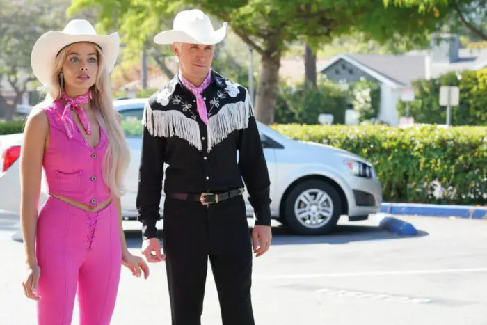 A man and woman in western wear stand on a sidewalk together in Barbie.