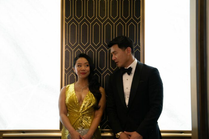 A woman in an elegant dress and a man in a tuxedo converse in an elevator.