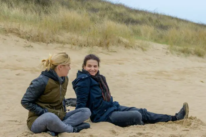 Marianne and Christèle sit together smiling on the beach, wearing jackets.