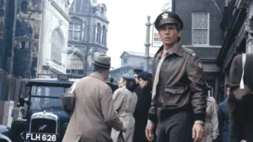 A man in uniform looks up a busy 1940s street.