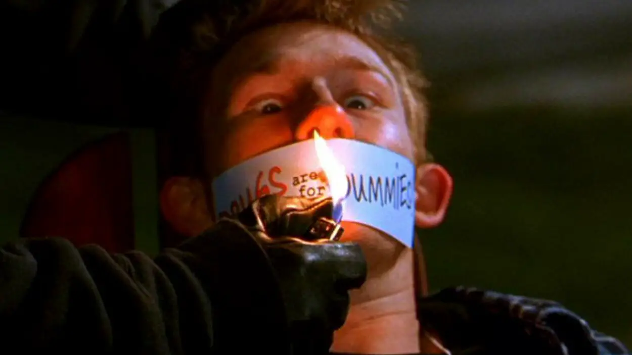A boy sits with tape over his mouth and someone wearing black gloves holding a lighter.