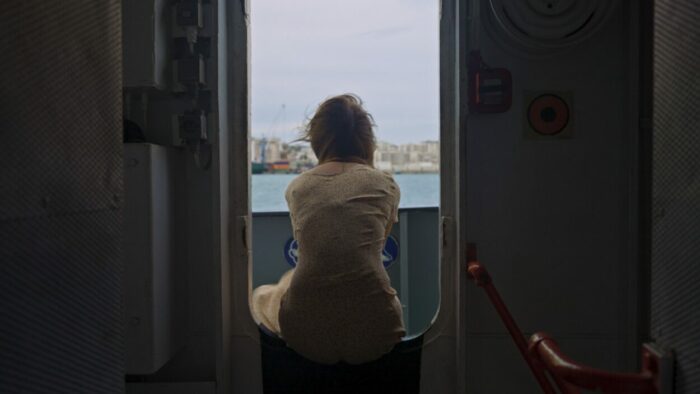 A woman sits in the doorway of a battleship, looking out at the harbor.
