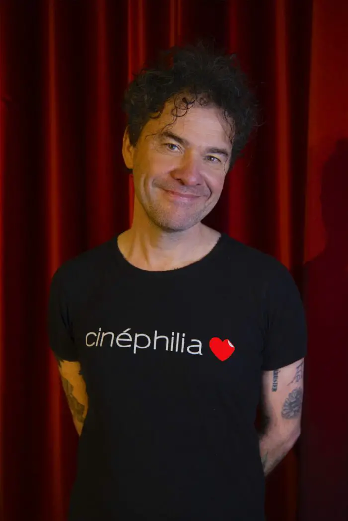 A publicity image of Mark Cousins wearing a black t-short with the lettering "Cinephelia"