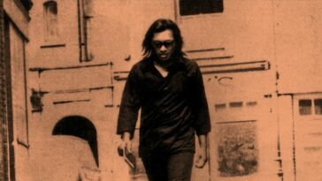 A man with long hair and sunglasses walks down the street.