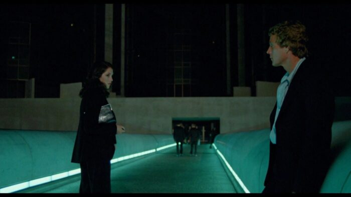 The Detective talks to The Player outside a neon-lit building.