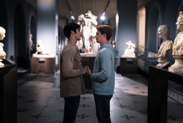 Alex and Henry stand center frame holding hands in a museum. 