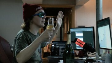 Keith Gill (Paul Dano) is live-streaming a YouTube video as YouTuber "Roaring Kitty" form his basement with sunglasses and a beanie on while he holds a glass of champagne.
