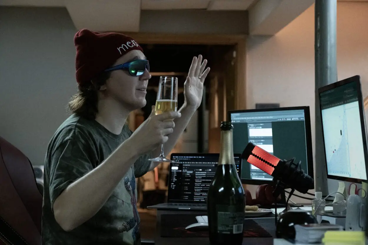 Keith Gill (Paul Dano) is live-streaming a YouTube video as YouTuber "Roaring Kitty" form his basement with sunglasses and a beanie on while he holds a glass of champagne.