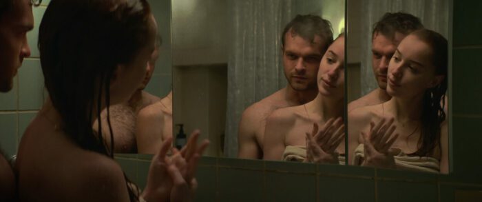 In an embrace, Luke and Emily look at a mirror after showering together while draped in towels. 