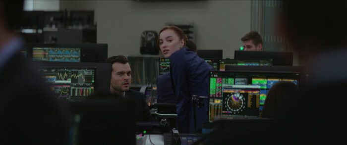 In a room filled with monitors displaying financial charts and graphs, Emily tells Luke a rumor she hears, changing their relationship as they both know it.