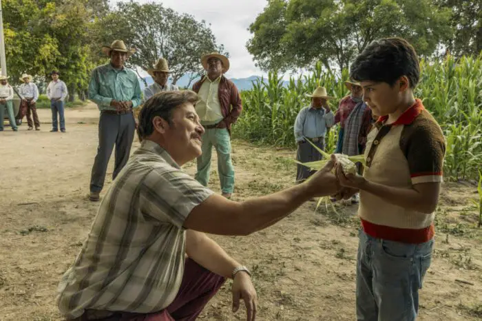 A kneeling father hands his son an ear of corn.