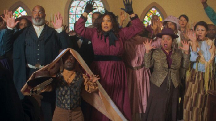 A small crowd stands with their hands up in a church in Outlaw Johnny Black