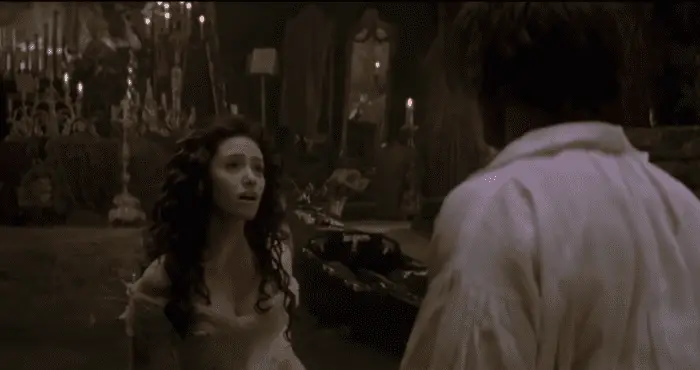 A young woman, with long brown hair and a white dress, looks at a man in front of her, there are lit candles behind her. 