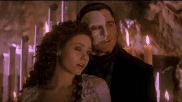 A man and a woman stand next to each other, her head on his shoulder. She is wearing a white dress and he has a white mask on half his face. Lit candles stand behind them.