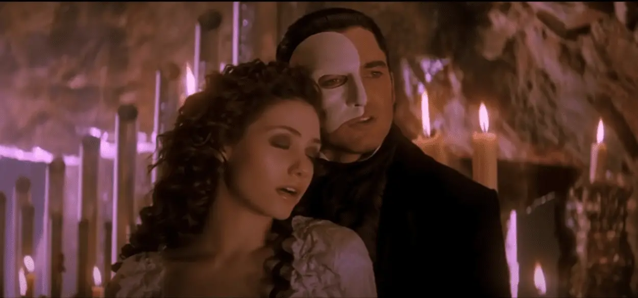 A man and a woman stand next to each other, her head on his shoulder. She is wearing a white dress and he has a white mask on half his face. Lit candles stand behind them.