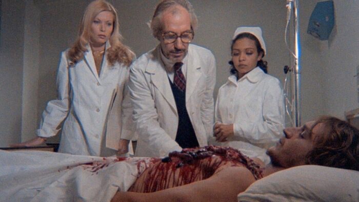 A doctor and two nurses look at a cadaver in bed whose heart has been ripped out of his chest.