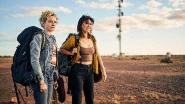 Julia Garner and Jessica Henwick star in The Royal Hotel, seen standing looking offscreen in the Australian Outback