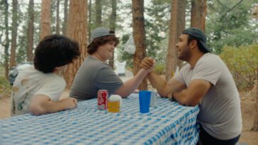 A teenage boy (Isaac Krasner) arm wrestles with his older cousin's boyfriend at a picnic table out in the wood
