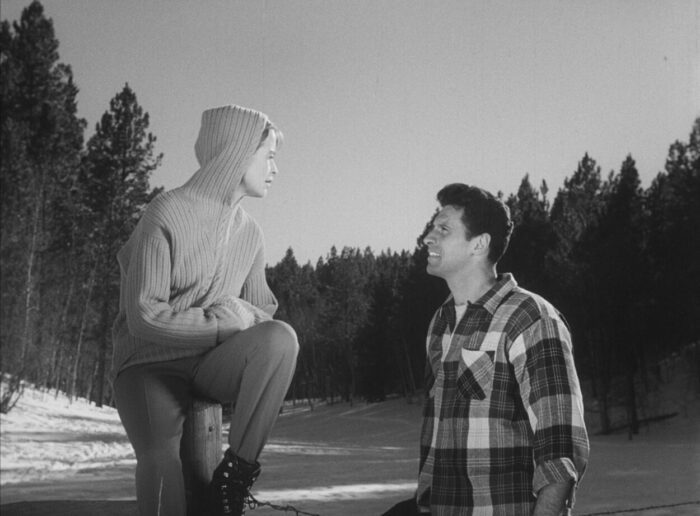 A man in flannel and a woman in a hoodie converse in a snowy forest.