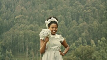Eva (Sandra Umulisa) stands against a verdant treeline dressed and garlanded in white flowers