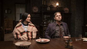 Mollie (Lily Gladstone) and Ernest (Leonardo DiCaprio) sit at a wood table with bowls and plated on the table. Ernest looks up and Mollie looks at him with admiration as she is draped with a knit shawl.