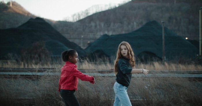 Two girls, one black and one white, walk in a field with a mountain range behind them.