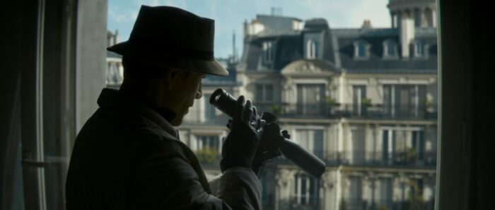 The Killer looks out a window with a telescope wearing a hat.