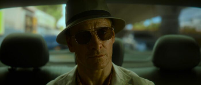 The Killer wears a hat and sunglasses in a car.