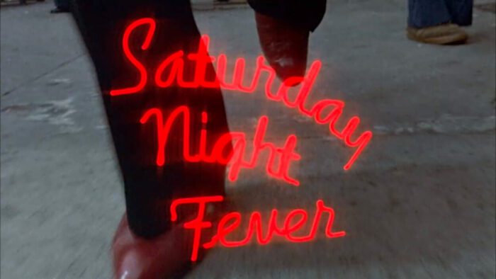 Title card of Satirday Night Fever, featuring those letters in neon red script as John Travolta's character walks down the street.