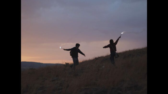 Two figures dance on a field in silhouette, each holding a small fire.