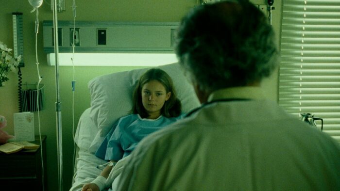 Cecilia lays in the hospital bed, staring at her doctor. There's a greenish hue in the room. 