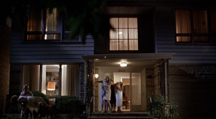 A darkly lit wide shot of Mrs. Lisbon on the front porch holding back her daughters from seeing the horrific sight of Cecilia, who is lying limp in her father's arms. 