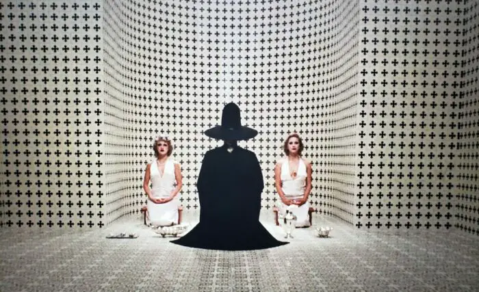 Connie de la Mora, Leticia Robles, and Alejandro Jodorowsky as the Bald women and the Alchemist in The Holy Mountain (1973), all three kneeling in a black and white room surrounded by strangely patterned walls, the alchemist all in black while the ladies wear white. Screen capture off Amazon.