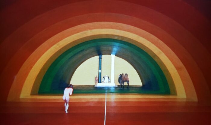 Horacio Salinas and Alejandro Jodorowsky as the Thief and the Alchemist in The Holy Mountain (1973). The Thief slowly approaches the Alchemist through a cavernous rainbow patterned hallway. Screen capture off Amazon.
