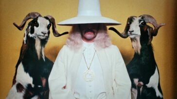 Alejandro Jodorowsky as the Alchemist in The Holy Mountain (1973). He sits dressed in white wearing a tall hat, flanked by taxidermy goats on their hindlegs. Screen capture off Amazon.