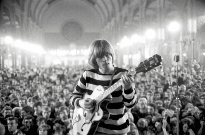 Brian Jones in THE STONES AND BRIAN JONES, facing the camera and playing guitar with a large crowd behind him.