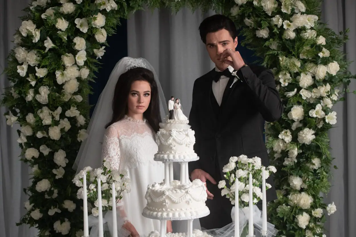 Priscilla (Caille Spaeny) and Elvis (Jacob Elordi) are cutting their wedding cake with Elvis smoking a cigarette and a white flower arch is in the background.