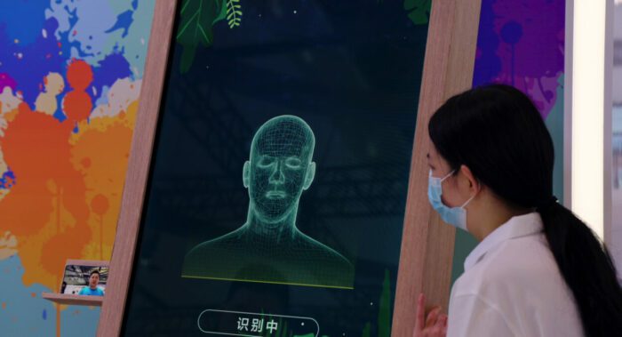 A Chinese woman wearing a mask looks at a computer screen displaying facial recognition. 