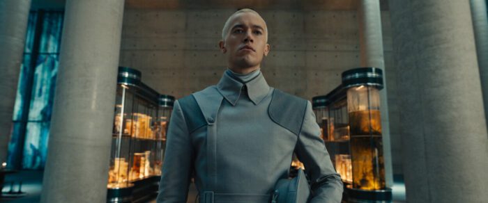 Coriolanus wears a gray Peacekeeper uniform with his new buzz cut as he prepares to work in District 12.