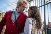 Coriolanus Snow (Tom Blythe), wearing a red vest outdoors, looks. passionately at Lucy Gray Baird (Rachel Zegler) behind gates, wearing something far less luxorious.