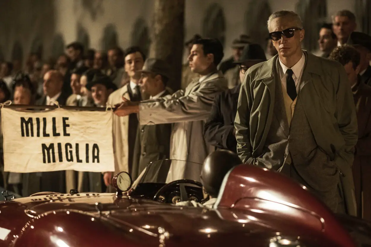 Adam Driver as Enzo Ferrari in Ferrari (2023). Photo credit Lorenzo Sisti. Owner EF Neon. Enzo Ferrari wearing sunglasses at night, standing beside one of his red racecars about to take off in the Mille Maglia race.