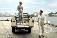 A woman stands on the back of a truck in a dress next to a man in a suit in Anyone But You