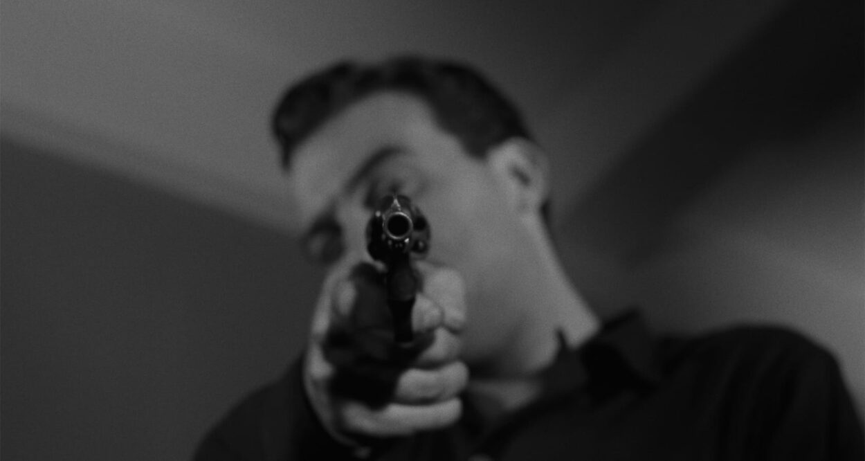 A short haired man aims a gun at the camera.