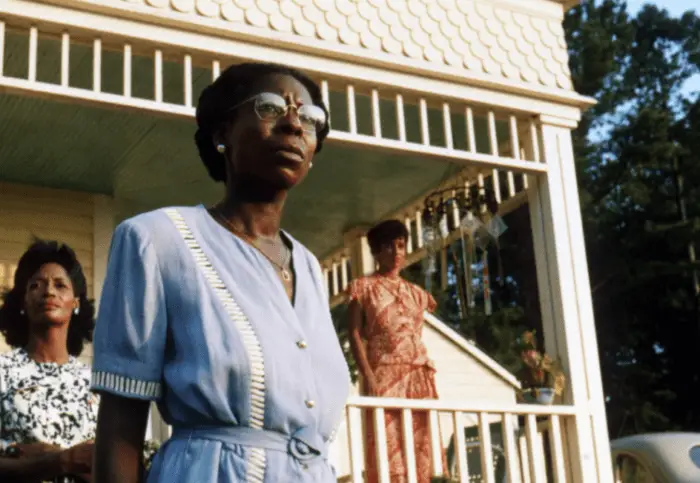 A woman wearing a purple dress and glasses stands in front of a porch in The Color Purple