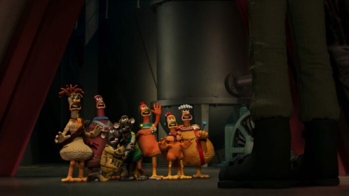 [L to R] Mac, Bunty, Nick, Fetcher, Ginger, Molly, and Babs voiced by Lynn Ferguson, Imelda Staunton, Romesh Ranganathan, Daniel Mays, Thandiwe Newton, Bella Ramsey, and Jane Horrocks in Chicken Run: Dawn of the Nugget (2023). Courtesy of Netflix. Most of the main chicken cast standing together as they try to flee the chicken nugget processing plant, their path obstructed by a human guard.