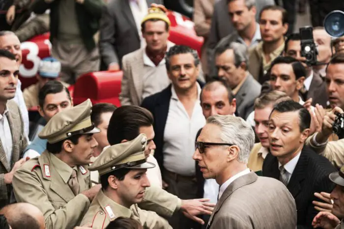 Adam Driver as Enzo Ferrari in Ferrari (2023). Photo credit Lorenzo Sisti. Owner EF Neon. A crowd clogs the street as Enzo Ferrari makes his way through, police clearing the way and reporters vying for attention.