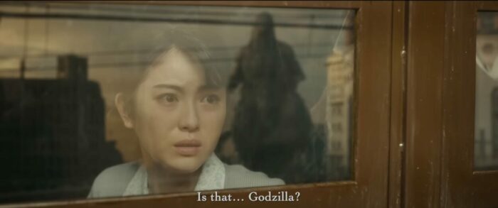Godzilla approaches a train, seen in the reflection of a window, as a woman on board stares in horror