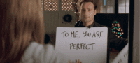 A man stands outside in a black shirt. He is holding a large white sign with the words "To me you are perfect" and a woman looks at him but we can only see her white shirt and brown hair