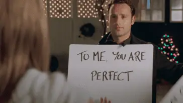 A man stands outside in a black shirt. He is holding a large white sign with the words "To me you are perfect" and a woman looks at him but we can only see her white shirt and brown hair